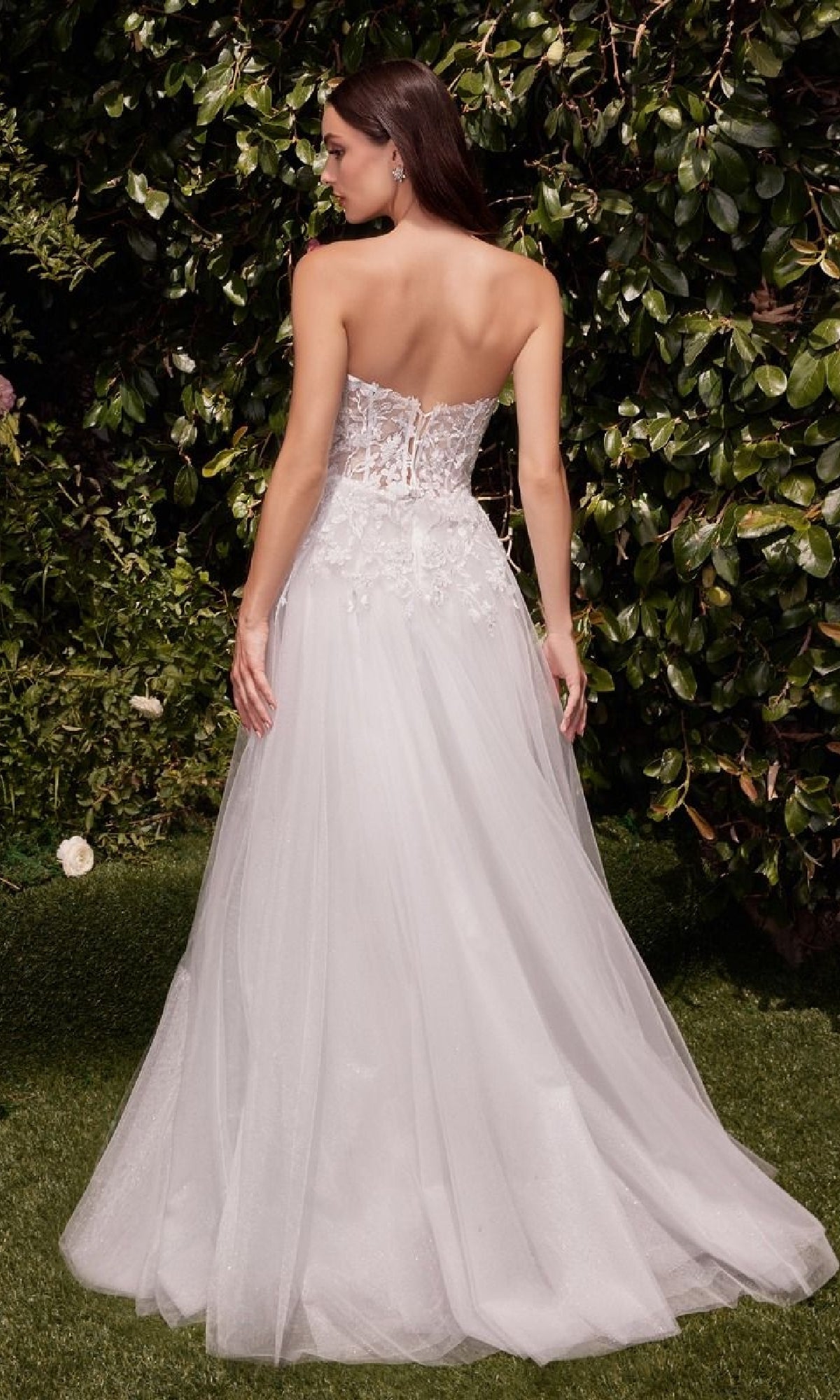 A woman stands outdoors with her back to the camera, surrounded by greenery, wearing the Strapless Long White Wedding Gown: Ladivine C148W featuring a lace corset bridal design.